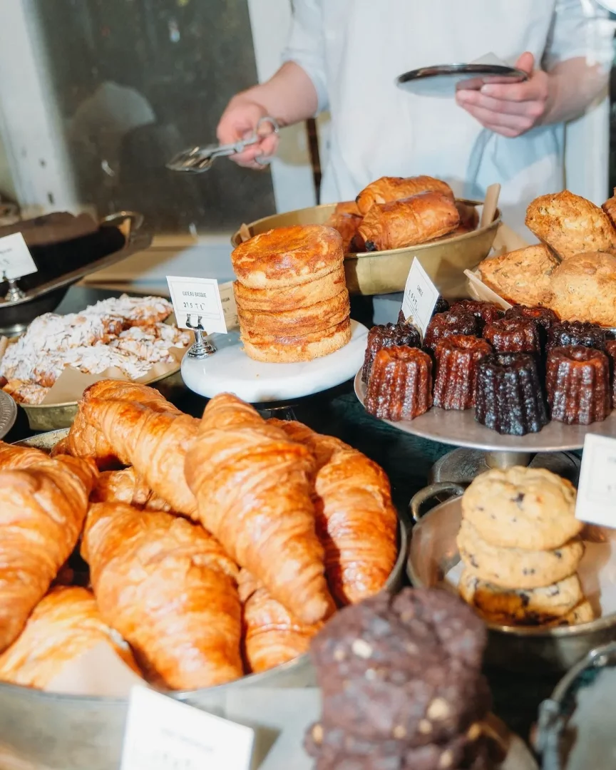 Petra Coffee'de sunulan göz alıcı taze hamur işleri vitrini; bol miktarda altın rengi kruvasan, çikolatalı caneléler, üst üste dizilmiş yuvarlak Gateau Basque ve kurabiyelerle dolup taşarken, arkada beyaz önlüklü bir çalışan titizlikle servis yapıyor.