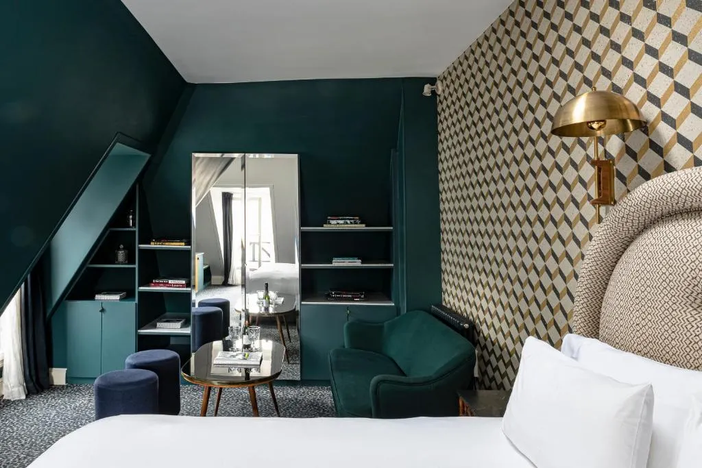 Koyu petrol mavisi duvarlar, geometrik desenli duvar kağıdı, rahat beyaz bir yatak ve aynalı bir dolabın yer aldığı modern ve şık bir otel odasında, koyu yeşil kadife bir koltuk, kitap dolu raflar ve üç küçük mavi tabure ile tamamlanmış zevkli bir tasarım görülüyor.