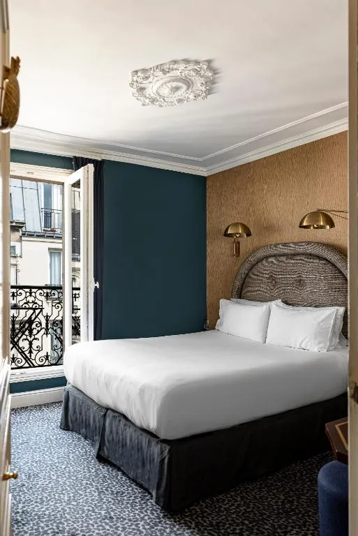 Pigalle bölgesindeki bu şık otel odasında, beyaz nevresimli yatak, koyu teal ve kahverengi duvarlar, süslü tavan rozeti ve Paris şehir manzaralı açık bir pencere modern ve klasik unsurları birleştiriyor.