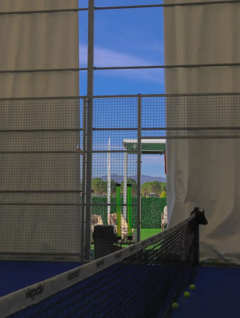 İzmir'deki bir padel kortunun içinden bakıldığında, mavi kort zemininde birkaç sarı topun yanında uzanan siyah file ağı ve tel örgü duvarların ötesinde yeşil ağaçlar, tepeler ve açık mavi gökyüzü görünen bir manzara.