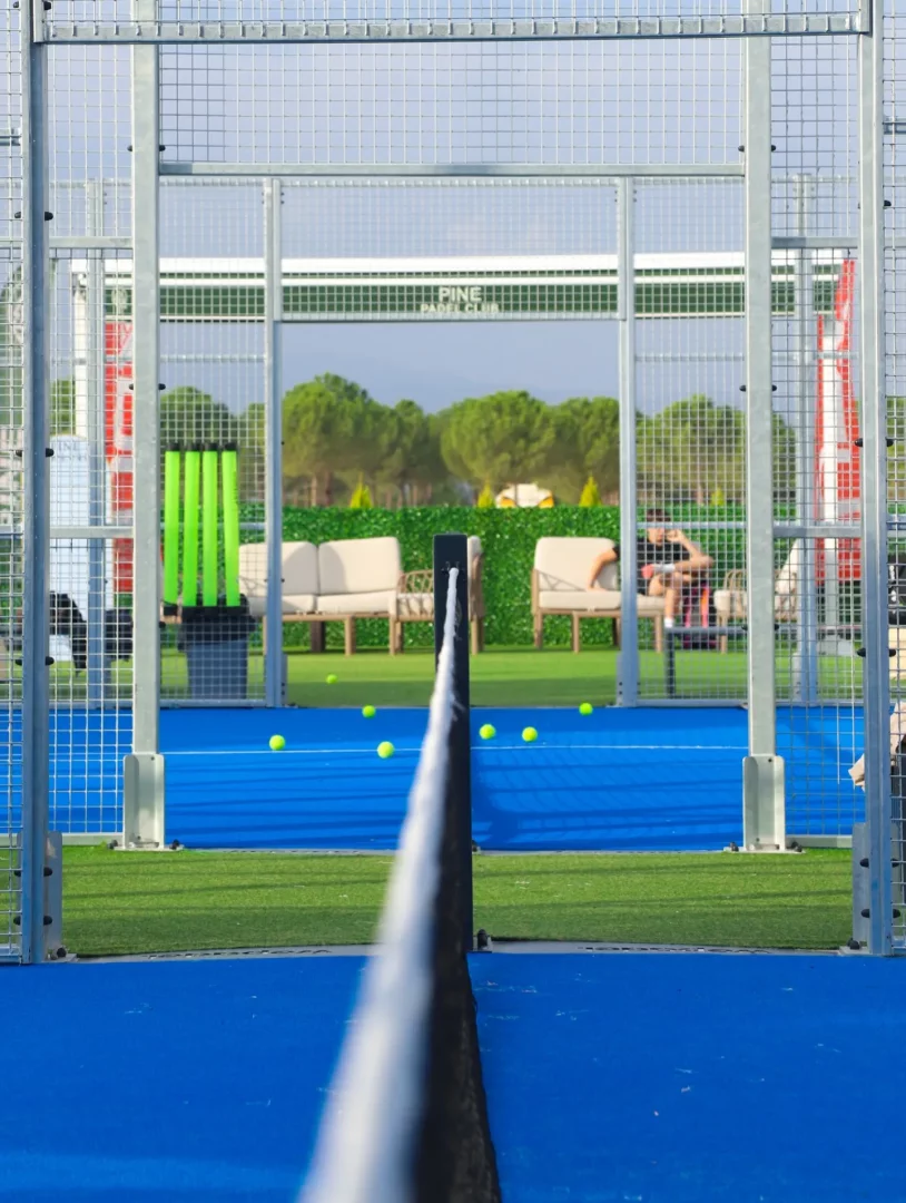 Pine Padel Club'ın İzmir'deki modern padel kortlarından birinin içinden çekilmiş bu fotoğrafta, parlak mavi oyun zemini, çevreleyen tel örgüler ve file belirginleşirken, arkada yeşil çam ağaçları ve dinlenme alanları yer alıyor.
