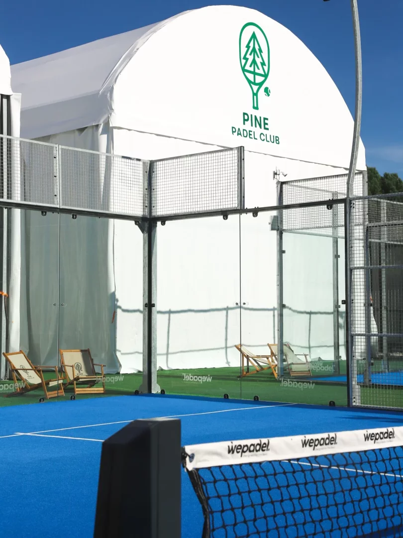 Mavi zeminli, fileli bir padel kortunun ön planda olduğu bu geniş açılı görselde, arkasında şeffaf cam duvarlar ve metal çitlerle çevrili modern bir padel kortu alanı, üzerinde yeşil çam ağacı logolu ve büyük harflerle PINE PADEL CLUB yazan beyaz, kemerli bir tesis binasıyla bütünleşiyor, dinlenme amaçlı şezlonglar da fark ediliyor.