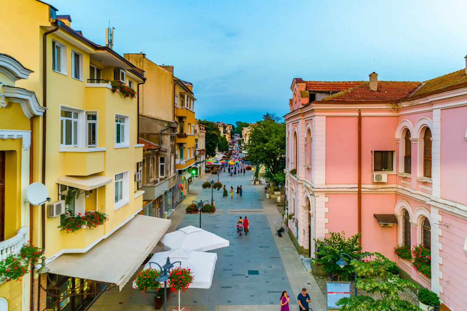 Mavi gökyüzü altında, sarı ve pembe tonlarındaki cephelere sahip tarihi binaların iki yanında yükseldiği, rengarenk çiçeklerle süslü lambaların ve şemsiyelerin bulunduğu, insanların rahatça dolaştığı hareketli bir yaya caddesi.