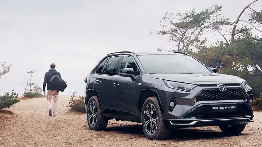 Kumlu bir patikada park etmiş koyu gri bir Toyota RAV4 Plug-in Hibrit SUV, ön planda net bir şekilde görünürken, arka planda ağaçlar ve sisli gökyüzü altında uzaklaşan bir kişi, plug-in hibrit araçların doğal ortamlardaki kullanımını vurguluyor.