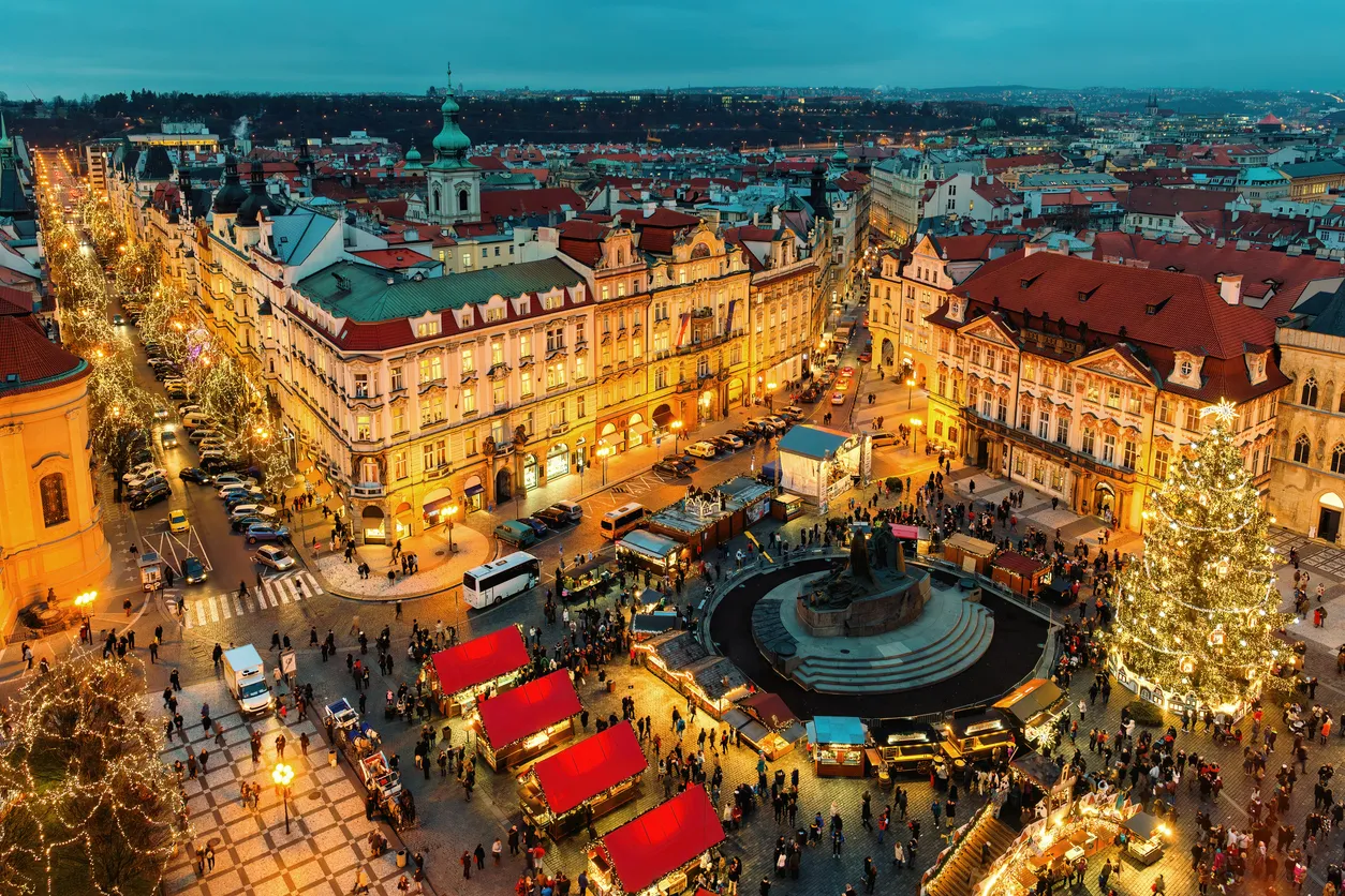 Prag'daki tarihi bir şehir meydanında, akşam vakti binlerce ışıkla bezenmiş devasa bir Noel ağacı ve kırmızı çatılı tezgahlarla dolu hareketli bir Noel pazarı, çevresindeki ışıl ışıl tarihi binalar ve caddelerle birlikte kalabalık bir kış atmosferi yaratıyor.