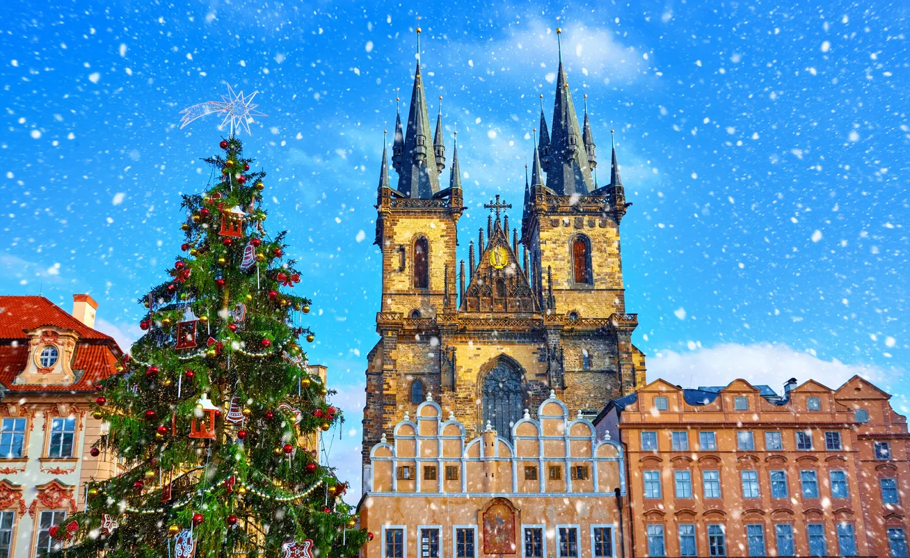 Prag'ın tarihi Eski Şehir Meydanı'nda Noel ruhunu yansıtan karlı bir kış gününde, altın, kırmızı ve fenerlerle süslenmiş ışıl ışıl bir Noel ağacı, Týn Kilisesi'nin görkemli gotik kuleleri ve çevresindeki tarihi binalarla birlikte etkileyici bir kartpostal görünümü sunuyor.