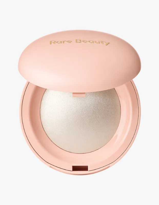 Açık pembe renkli, üst kapağında altın rengi Rare Beauty yazısı yer alan ve içinde ışıltılı, gümüş tonlarında Positive Light Silky Touch aydınlatıcının göründüğü, hafifçe aralanmış kompakt bir makyaj ürününün yakından görünümü.