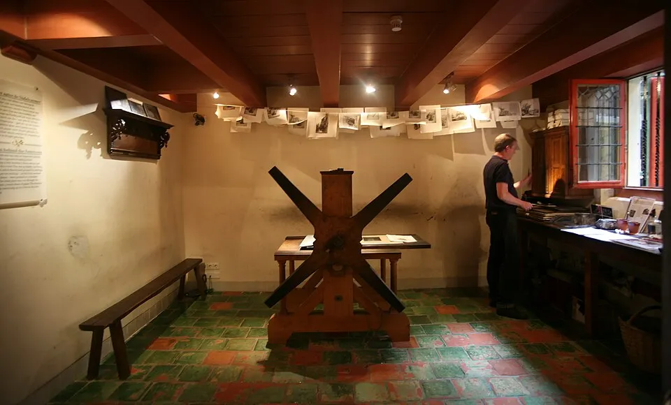 Rembrandt Evi'nin tarihi atmosferini yansıtan bu iç mekanda, ahşap tavan kirişlerinin altında, odanın ortasında çapraz kollu büyük bir ahşap baskı presi yer alırken, duvarlarda sergilenen küçük kağıtlar ve sağda duran bir ziyaretçi dikkat çekiyor.