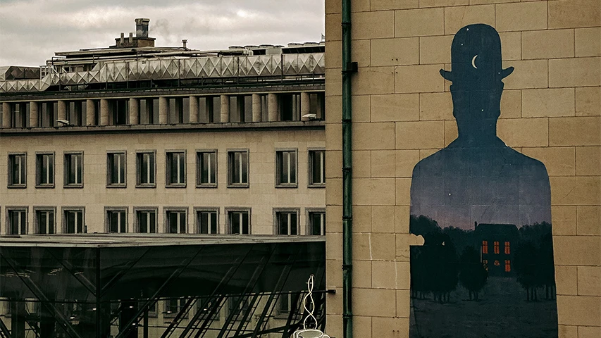 Sağdaki tuğla duvarda, René Magritte'in sanat anlayışına gönderme yapan, melon şapkalı bir adamın içinde ay ve yıldızların olduğu gece gökyüzü ile ışıklı pencereleri olan bir evin ve ağaçların bulunduğu kırsal manzarayı içeren siluetinin yer aldığı büyük bir duvar resmi görülmekte; bu sürrealist görüntü, sol taraftaki pencereli modern bir binanın ve bulutlu bir gökyüzünün önünde durmaktadır.