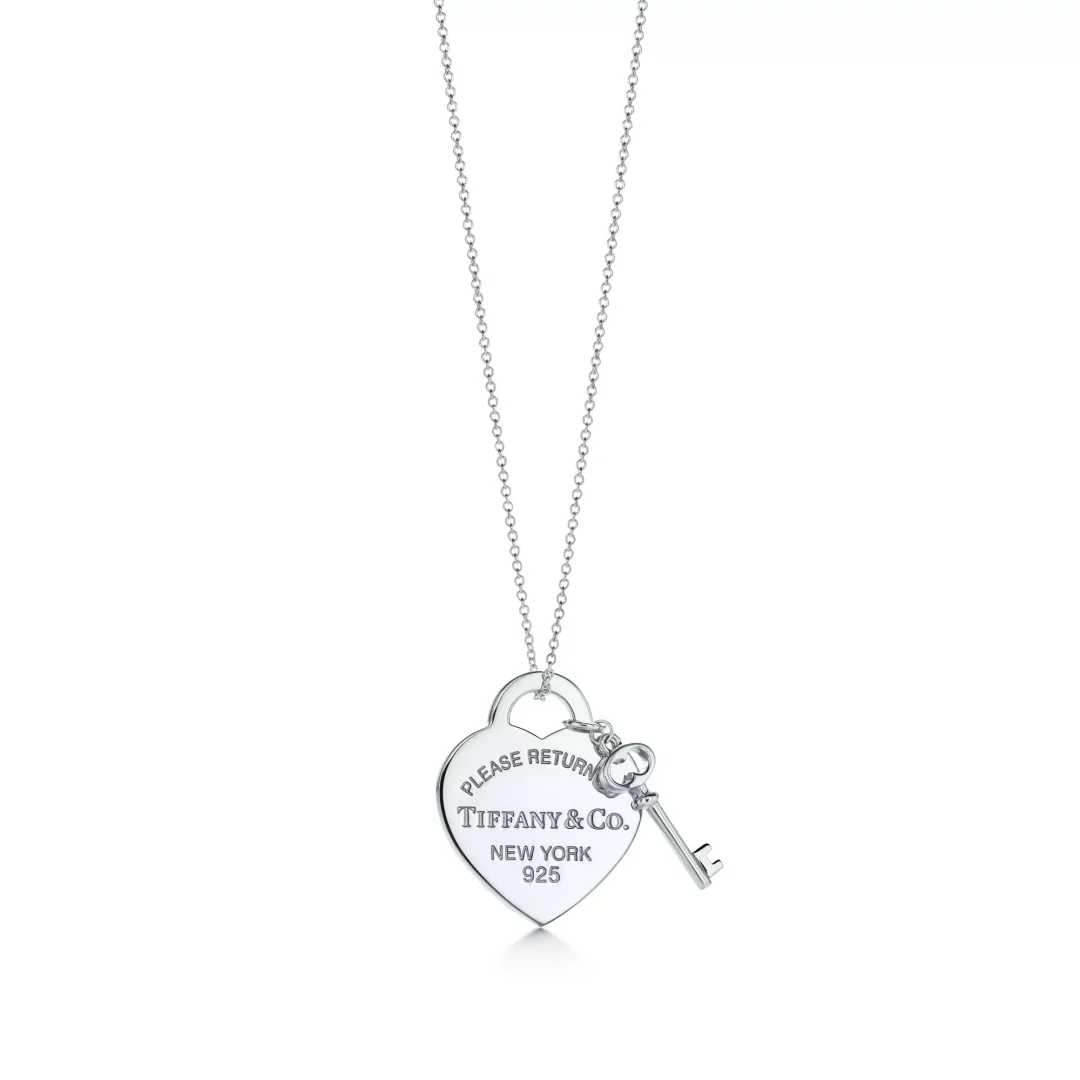 Bu görsel, Tiffany & Co.'nun Return to Tiffany koleksiyonu temasına uygun, üzerinde PLEASE RETURN TIFFANY & Co. NEW YORK 925 yazısı bulunan parlak gümüş kalp kolye ucu ve ona takılı zarif bir anahtardan oluşan sade zincirli bir kolyeyi beyaz bir zeminde sergiliyor.