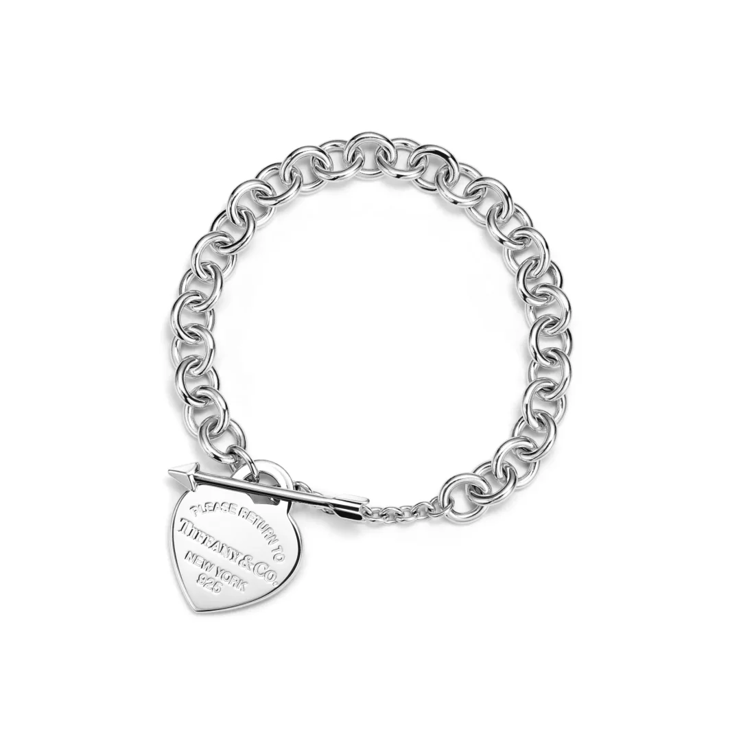 Beyaz bir arka plan üzerinde, üzerinde PLEASE RETURN TO TIFFANY & CO. NEW YORK 925 yazısı bulunan kalp şekilli bir madalyonun T-bar klips ile bağlı olduğu parlak gümüş zincir bileklik, ikonik Return to Tiffany koleksiyonunun dikkat çekici bir parçasını temsil ediyor.