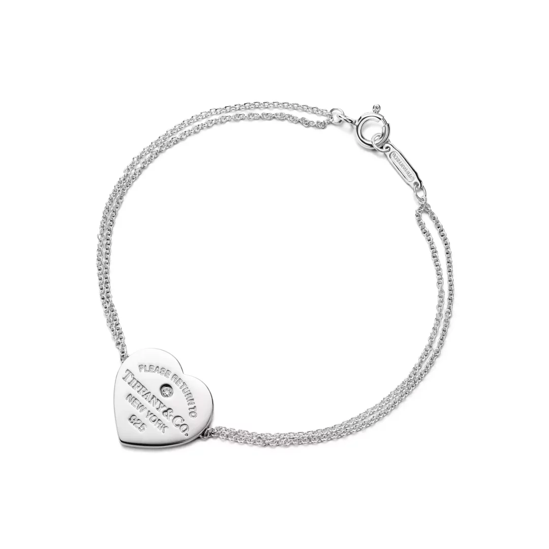 Beyaz arka planda sergilenen, çift zincirli, gümüş rengi Return to Tiffany koleksiyonu bileklik, ortasında pırlanta işlemeli ve üzerinde PLEASE RETURN TO TIFFANY & CO. NEW YORK 925 yazan parlak kalp şeklinde bir charm ile özenle tasarlanmıştır.