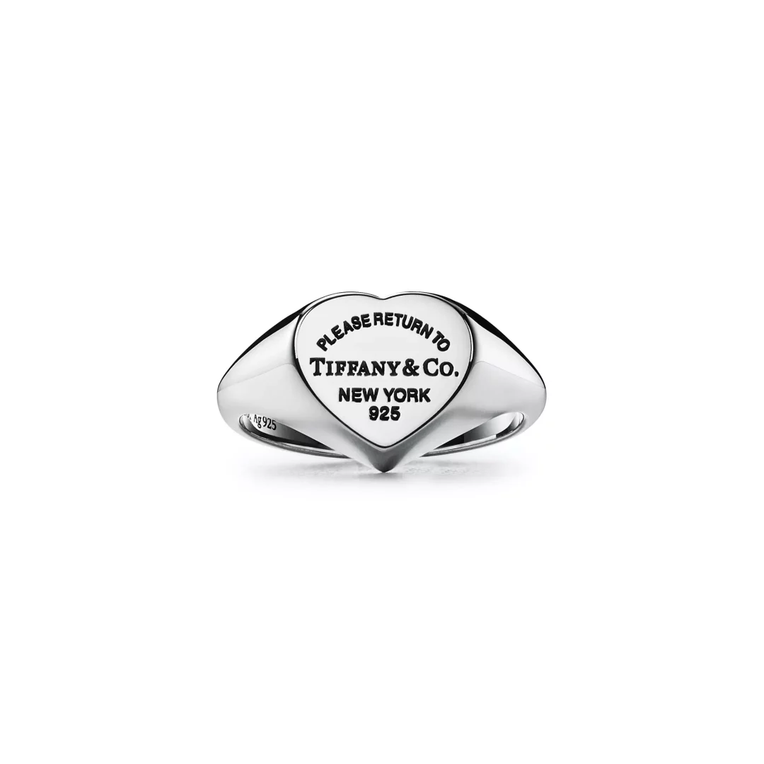 Gümüş renkli, kalp şeklinde üst yüzeye sahip bir mühür yüzüğü, üzerinde PLEASE RETURN TO TIFFANY & CO. NEW YORK 925 yazısı oyulmuş ve bu özel tasarım Return to Tiffany koleksiyonunun ikonik bir parçası olarak beyaz bir arka plan üzerinde sergileniyor.