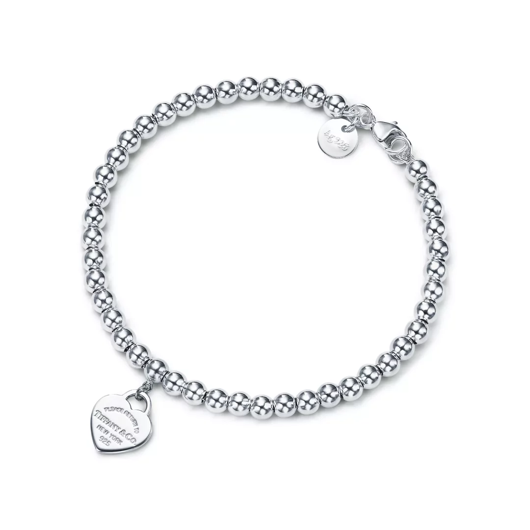 Return to Tiffany Koleksiyonu'na ait, parlak gümüş rengi yuvarlak boncuklarla dizilmiş, üzerinde Tiffany & Co. yazılı kalp şeklinde bir etiket ve Ag 925 damgalı küçük yuvarlak bir charm bulunan zarif bir gümüş bileklik beyaz zeminde şık bir şekilde sergileniyor.