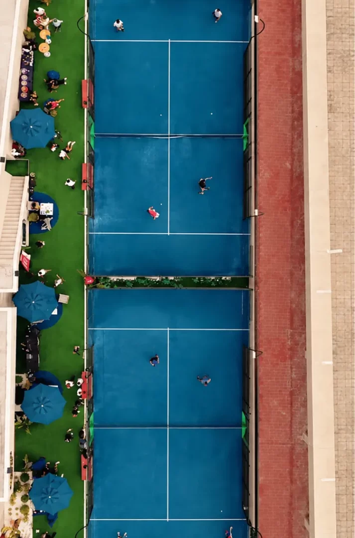 Rixos Tersane Padel Kulübü'nün havadan çekilmiş bu görüntüsünde, mavi zeminli çok sayıda padel kortunda raketleriyle aktif olarak oynayan oyuncular ve yanlarındaki yeşil dinlenme alanında şemsiyelerin altında oturan insanlar dikkat çekiyor.