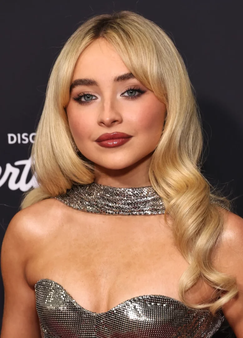 Sabrina Carpenter'ın omuzlarına dökülen sarı, dalgalı saçları ve yüzünü çevreleyen modern perçem modeli ile göz kamaştırıcı makyajının vurgulandığı, gümüş rengi parıltılı straplez elbisesi içinde çekilmiş yakın plan bir portresi.