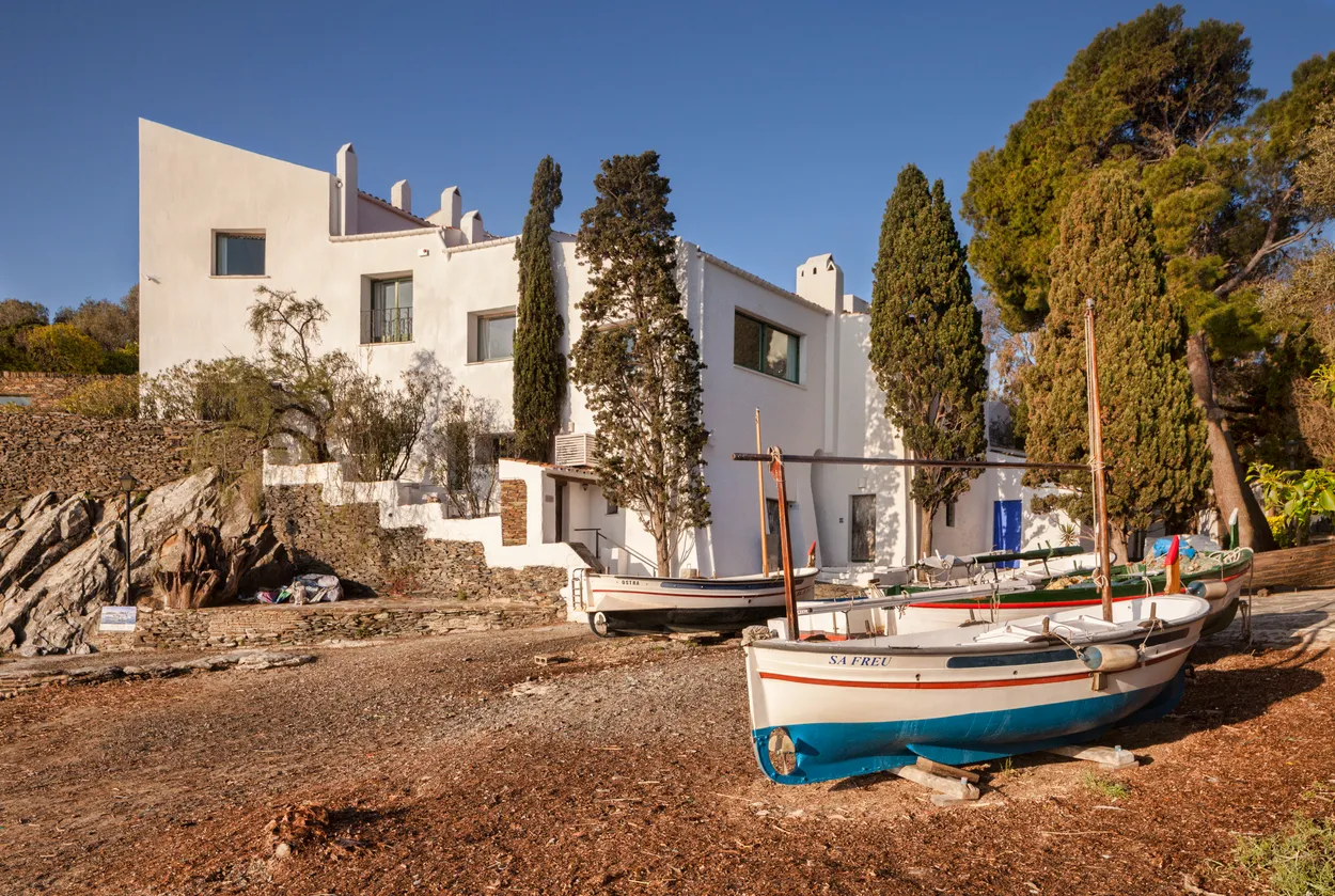 İspanya'nın Portlligat koyunda yer alan Salvador Dalí'nin Evi, beyaz badanalı modernist mimarisiyle kayalık bir yamaçta yükselirken, ön plandaki çakıllı kıyıda çeşitli ahşap tekneler güneşin altında dinleniyor, Akdeniz'in huzurlu atmosferini gözler önüne seriyor.