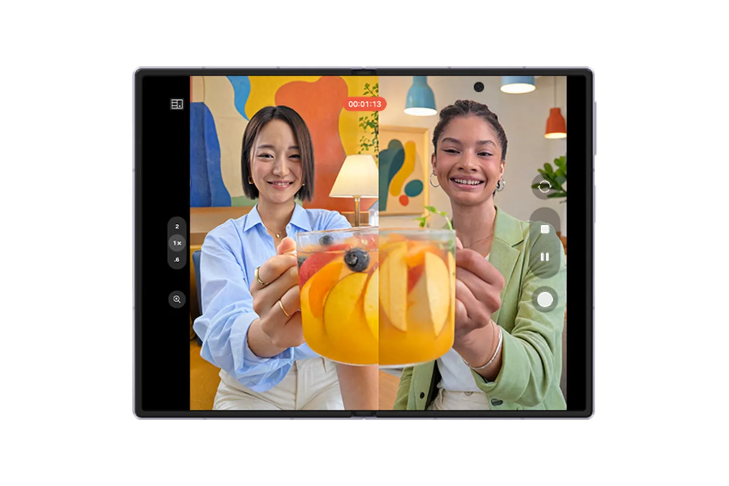 Samsung Galaxy Z Fold benzeri bir katlanabilir telefonun büyük ekranında, iki farklı kadının meyve dilimli içeceklerini kameraya doğru uzatırken gülümsediği, bölünmüş ekran görüntüsüyle aynı anda çekim veya görüntülü arama yeteneğini sergileyen canlı bir video kaydı.
