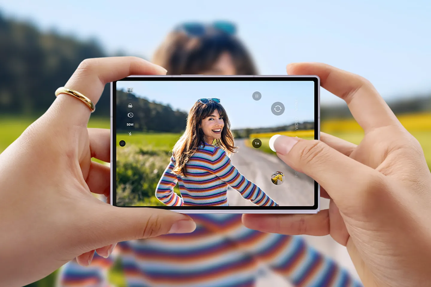 Güneşli yeşil bir dış mekanda neşeyle kameraya gülümseyen, renkli çizgili süveterli genç bir kadını gösteren ve 50M kamera ayarı ile fotoğraf arayüzünü sergileyen bir Samsung Galaxy Z Fold telefon ekranını yatay tutan eller.
