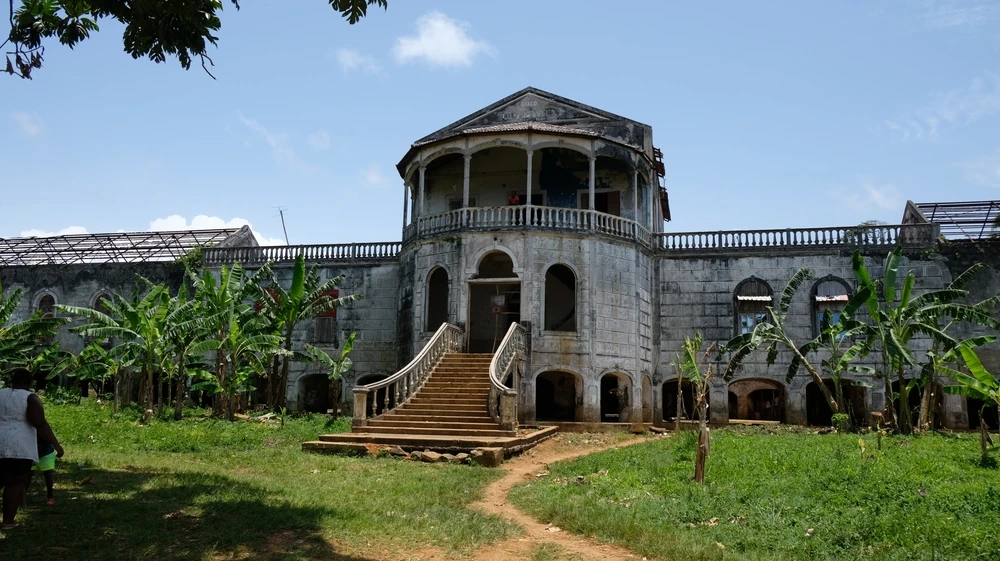 São Tomé ve Príncipe'nin UNESCO Dünya Mirası Listesi'ne eklenen roça'larından biri olan bu görselde, çevresi muz ağaçlarıyla sarılı, iki katlı, merdivenli girişi ve geniş balkonuyla dikkat çeken, Portekiz kolonyal dönemine ait, bakımsız ancak çarpıcı bir plantasyon binası gün ışığında görünüyor.