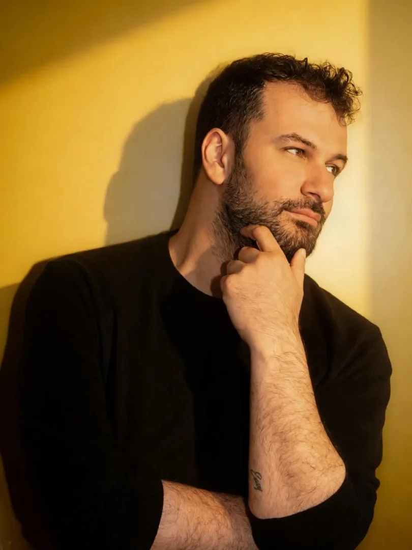 Selim Can Çelik'in yakın çekim portresinde, düşünceli bir ifadeyle sağ üst köşeye doğru bakarken, kıvırcık saçları ve gür sakalıyla çenesini eline dayadığı, sol ön kolunda belirgin bir dövme bulunan siyah uzun kollu tişörtüyle sıcak sarı bir duvar önünde aydınlık bir ışıkla öne çıktığı görülüyor.