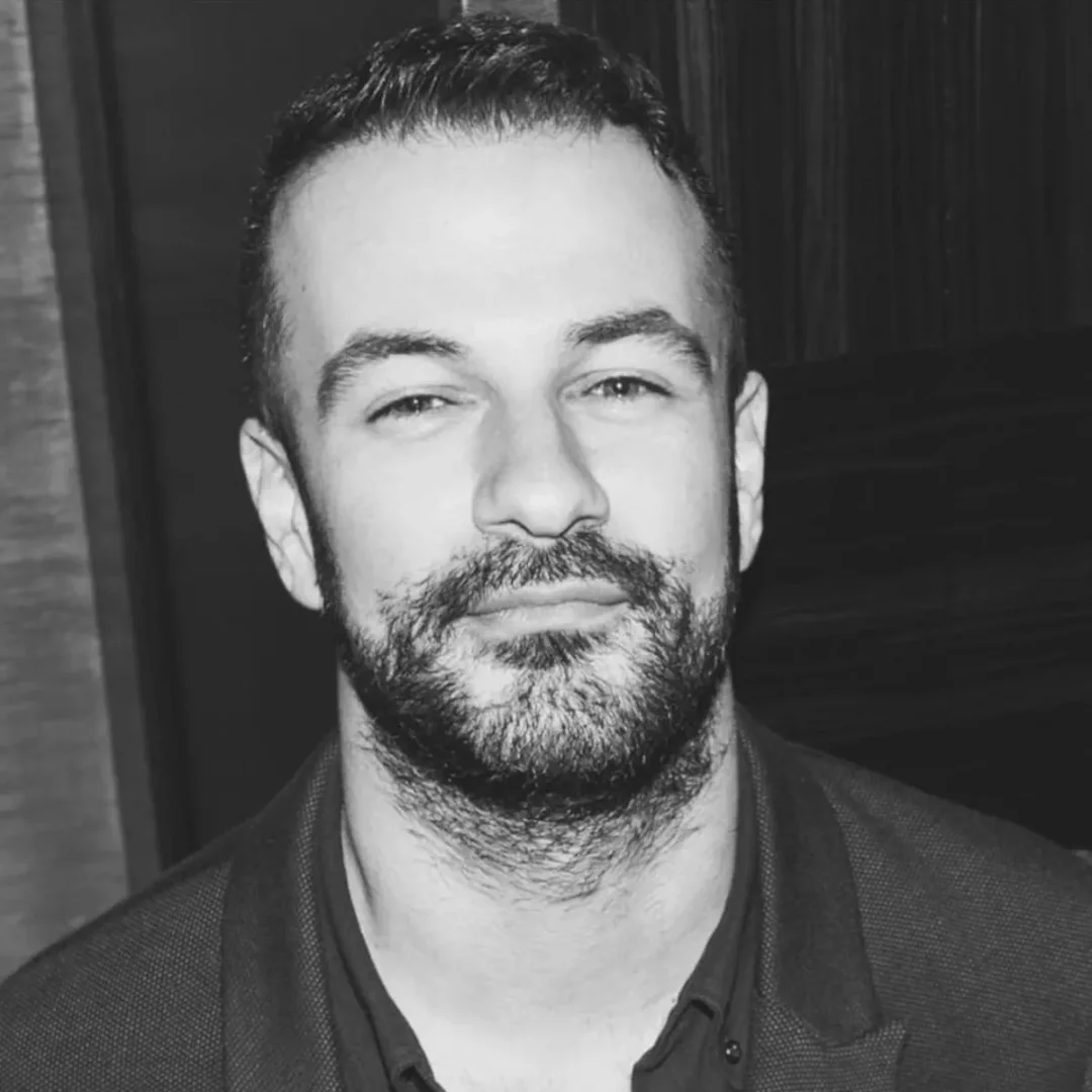 Selim Can Çelik'in koyu renk saçlı, sakallı, hafif ciddi bir ifadeyle doğrudan kameraya baktığı, omuz hizasından yukarı çekilmiş siyah beyaz portresi, karanlık ve bulanık bir arka planın önünde belirgin bir şekilde duruyor.