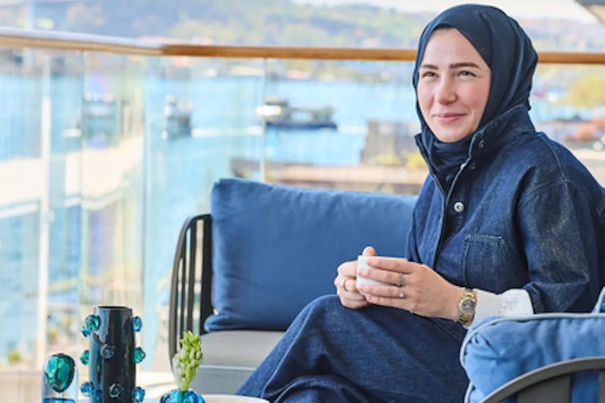 Seym Glass kurucusu Şeyma Nur Soysal Karpuz, lacivert başörtüsü ve denim ceketiyle, iki eliyle beyaz bir kupa tutarken, solunda sergilenen eşsiz el işçiliği cam tasarımlarının ve arkasındaki Bosphorus manzarasının eşlik ettiği balkonda oturmuş gülümsüyor.