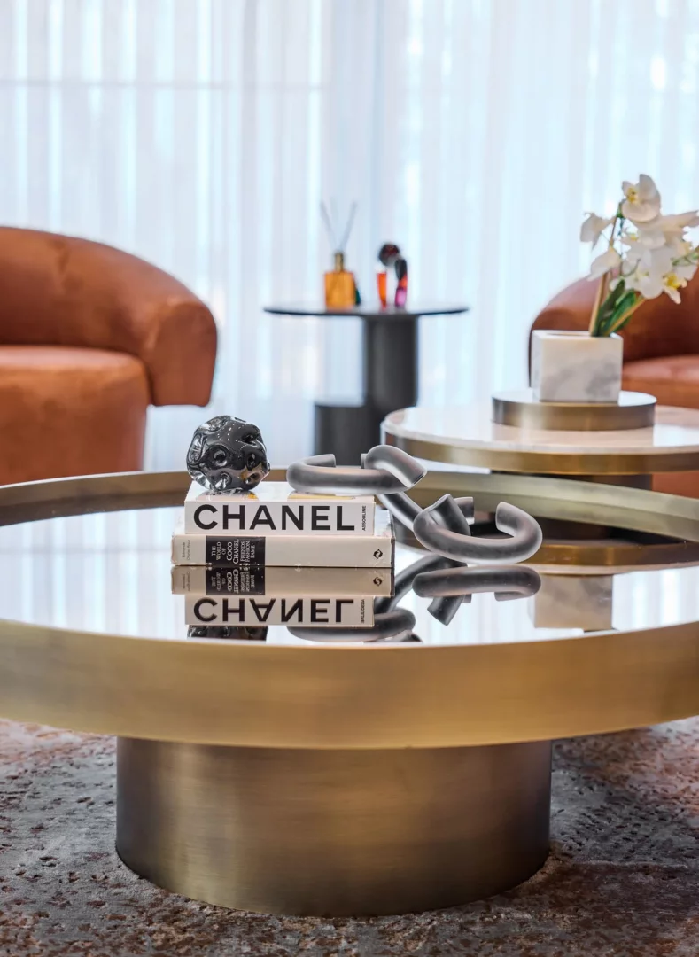 Bir lüks salon sehpası üzerinde, ayna yüzeyine yansıyan Chanel kitapları, koyu renkli oyma detaylı küresel bir cam obje ve soyut kavisli cam objelerle şık bir düzenleme sergilenirken, arka planda modern koltuklar ve zarif dekoratif detaylar bulanık bir şekilde görülüyor.