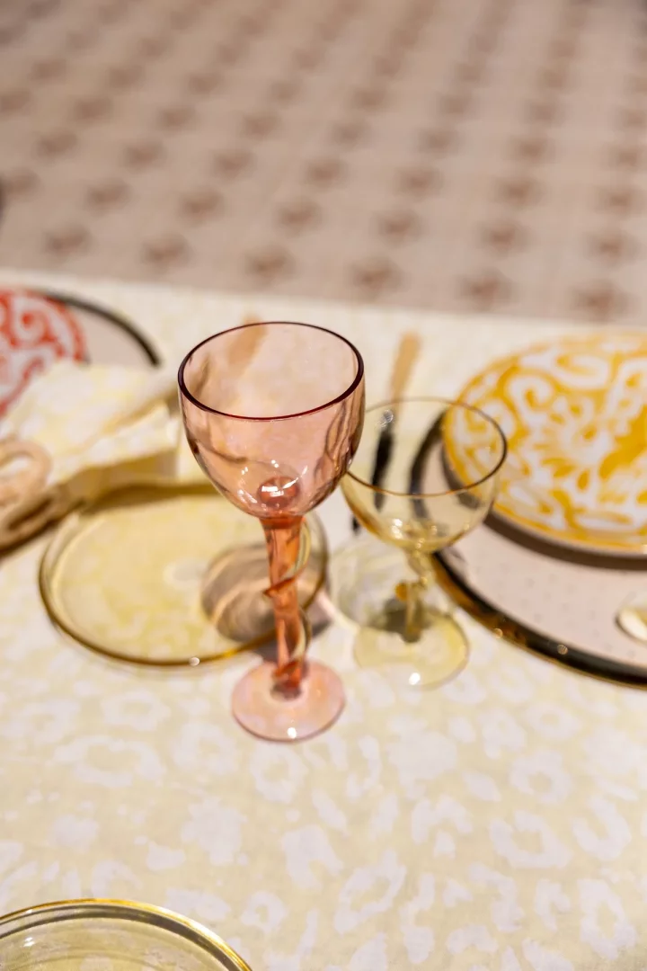 Şık bir masa düzeninde, ön planda belirgin burgulu sapı olan pembe kadeh ile arka planda aynı zarif tasarıma sahip sarı kadeh, özel anlar için tasarlanmış estetik Şeym Glass ürünlerini sergiliyor.