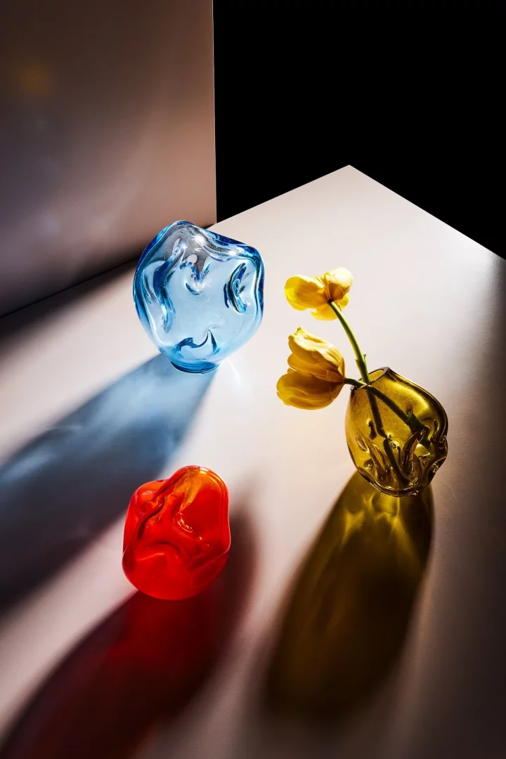 Bir stüdyo çekiminde, Seym Glass'ın camın hikâyesini ve el işçiliğini vurgulayan, ışıkla etkileşimi zenginleştiren, organik formlara sahip, mavi, turuncu-kırmızı ve sarı-yeşil renklerdeki üç el yapımı cam obje, güçlü gölgeleriyle birlikte estetik bir kompozisyon oluşturuyor.
