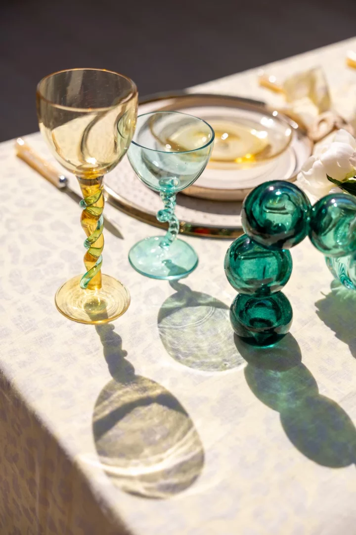 Şeyma Nur Soysal Karpuz'un kurucusu olduğu Seym Glass markasının özgün el işçiliği ve camın ışıkla kurduğu büyüleyici ilişkiyi gözler önüne seren bu sofra düzeninde, bükümlü saplarıyla dikkat çeken sarı ve turkuaz renkli cam kadehler ile üçlü yeşil dekoratif cam küreler, masa örtüsü üzerinde uzun ve belirgin gölgelerle camın hikayesini çağrıştırıyor.