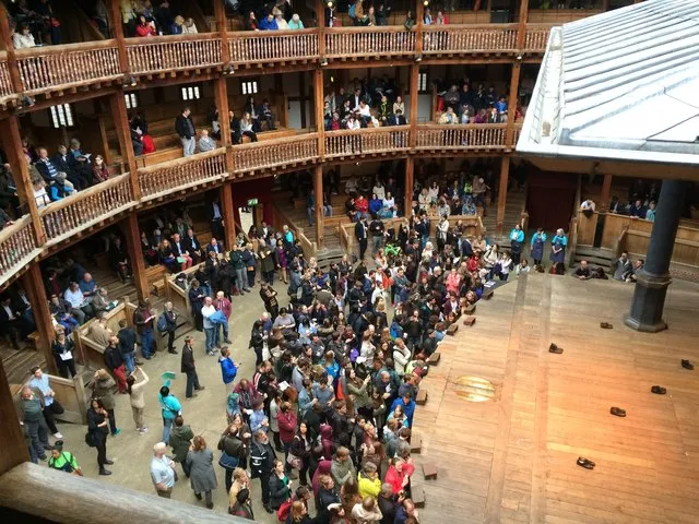 Shakespeare’s Globe açık hava tiyatrosunun ahşap galerilerinde ve ortadaki geniş avlusunda toplanmış, Elizabeth dönemi tiyatro anlayışını yansıtan kalabalık bir izleyici kitlesinin, canlı bir etkinlik veya atölye programına katılımını gösteren genel bir iç görünüm.