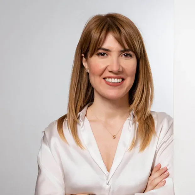 Sinem Olcay, omuzlarına dökülen kahverengi saçları ve kahkülleriyle neşeli bir şekilde gülümserken, beyaz yakalı saten bir bluz giymiş ve kollarını önünde kavuşturmuş bir poz veriyor; boynunda zarif, küçük bir altın kolye ve kulaklarında küpeler dikkat çekiyor, açık renk arka plan önünde aydınlık ve profesyonel bir portre sunuyor.