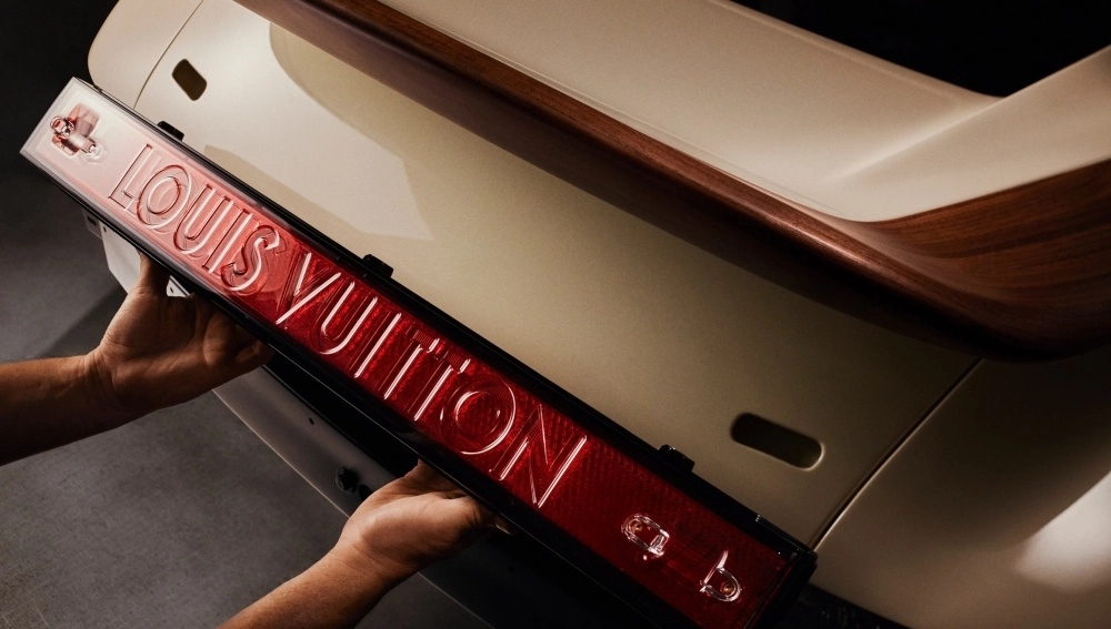 Ellerinde tutulan, Louis Vuitton yazılı, kırmızı renkli özel üretim arka panel, bej rengi ve ahşap detaylı bir Porsche 911 restomod aracın arka kısmına titizlikle yerleştirilirken, bu özel iş birliğinin zanaatkarlığını vurguluyor.