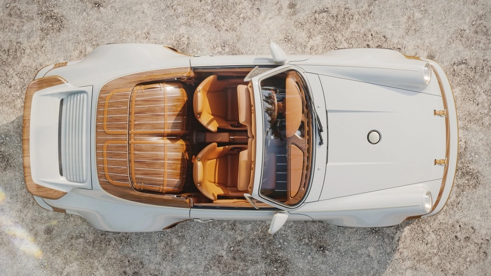 Beyaz renkli, üstü açık bir Singer Porsche 911 restomod aracının drone çekimi, kahverengi deri koltukları ve arka bölümünü süsleyen zarif ahşap panellerle zenginleşen, lüks bir iş birliğinin estetiğini vurgulayan özel bir koleksiyon parçasını sergiliyor.
