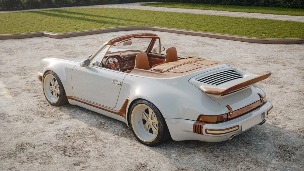 Açık gri renkte, üstü açık, kahverengi deri iç mekana ve ahşap detaylarla zenginleştirilmiş Singer tarzı bir restomod Porsche 911 cabrio, arka sağ çaprazdan kumlu zeminde güneşin altında park edilmiş duruyor, klasik ve modernin lüks bir birleşimini sergiliyor.