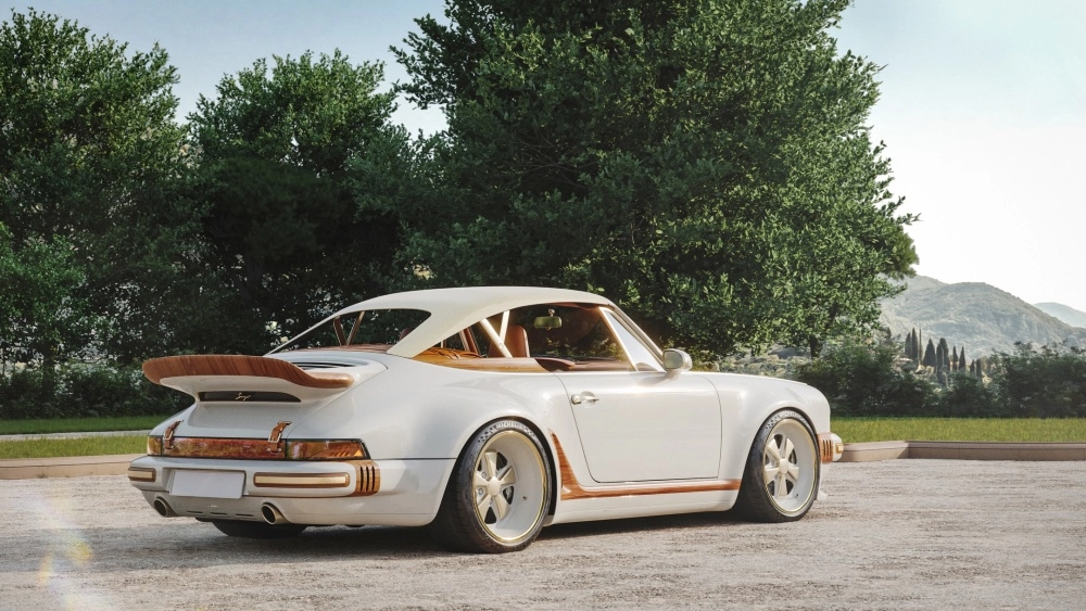 Beyaz renkli, arkadan hafif açılı görünen bu Singer Porsche 911 restomod, yükseltilmiş açık renkli tentesi, dikkat çekici ahşap spoyleri ve yan şeritleriyle, yeşil ağaçların ve dağların çevrelediği çakıllı bir alanda zarifçe sergileniyor.