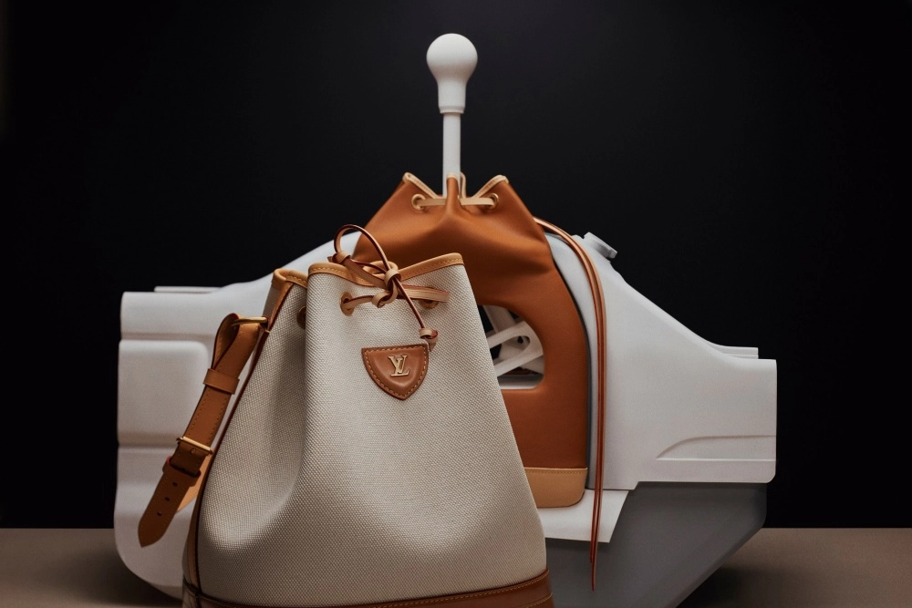 Ön planda, beyaz vites kolu olan bir otomobil konsolu benzeri yapının üzerinde, Louis Vuitton logolu krem rengi kanvas ve tann rengi deri detaylı bir kova çanta ile arkasında görünen benzer bir deri çanta, Singer ve Louis Vuitton iş birliğinin Porsche 911 restomod temalı deri koleksiyonundan parçaları sunuyor.