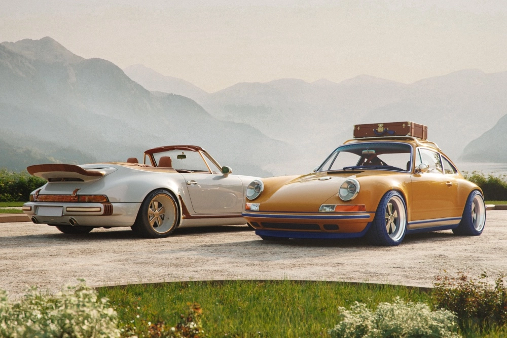 Puslu dağların önünde, açık bej rengi üstü açık ve hardal sarısı kapalı olmak üzere iki özel Singer Porsche 911 restomod otomobili, zarif dış tasarım detaylarını sergileyerek bir iş birliği koleksiyonundan çarpıcı bir sahne sunuyor; sarı olanın tavanında klasik bir bavul bulunuyor.