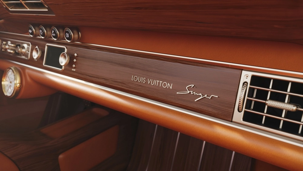 Singer ve Louis Vuitton iş birliğiyle hazırlanan bir Porsche 911 restomod aracın iç mekanından detay: Torpido boyunca uzanan koyu kahve ahşap panelde Louis Vuitton ve Singer logoları ile taba rengi deri döşemeler, klasik göstergeler ve krom detaylı havalandırma ızgarası, lüks ahşap işçiliğini vurguluyor.