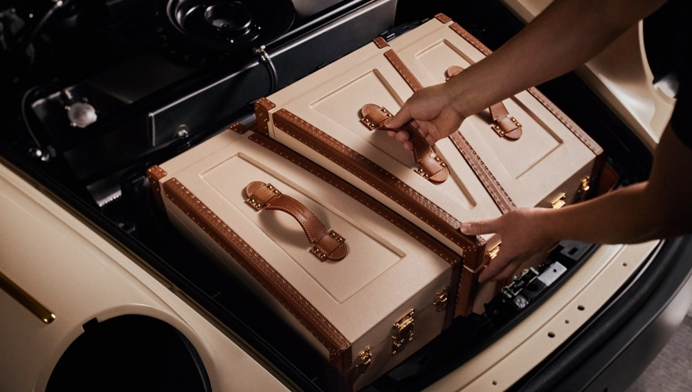 Bir kişinin elleri, Singer Porsche 911 restomod için Louis Vuitton ile yapılan özel iş birliği kapsamında, krem rengi gövdesi ve kahverengi deri detayları ile özenle tasarlanmış iki adet şık valizi veya sandığı aracın ön bagajına dikkatlice yerleştirirken görülüyor.
