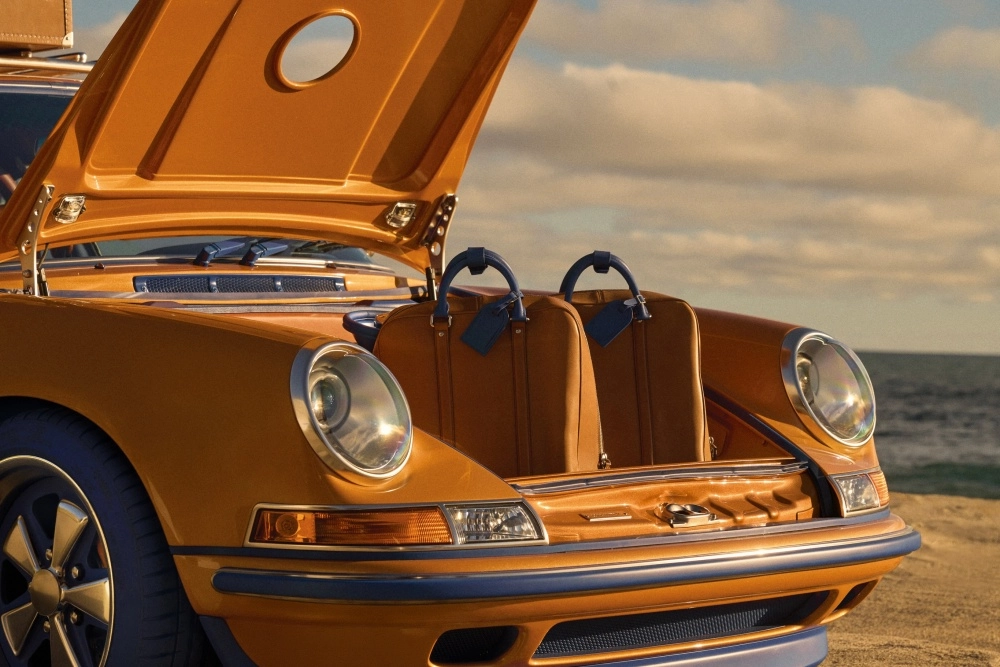 Sahilde duran altın sarısı, mavi detaylı bir Singer restomod Porsche 911'in ön bagaj kapağı açık bir şekilde görülüyor, bagajda mavi kulplu özel tasarım iki kahverengi Louis Vuitton valiz dikkat çekiyor.