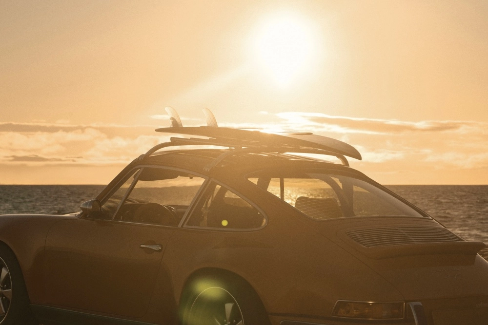 Gün batımının altın rengi ışıkları altında denizin kenarında duran, tavanında bir sörf tahtası taşıyan klasik bir Porsche 911 otomobili, Singer'ın restomod iş birliği ruhunu yansıtan estetiğiyle sıcak ve özgür bir anı betimliyor.