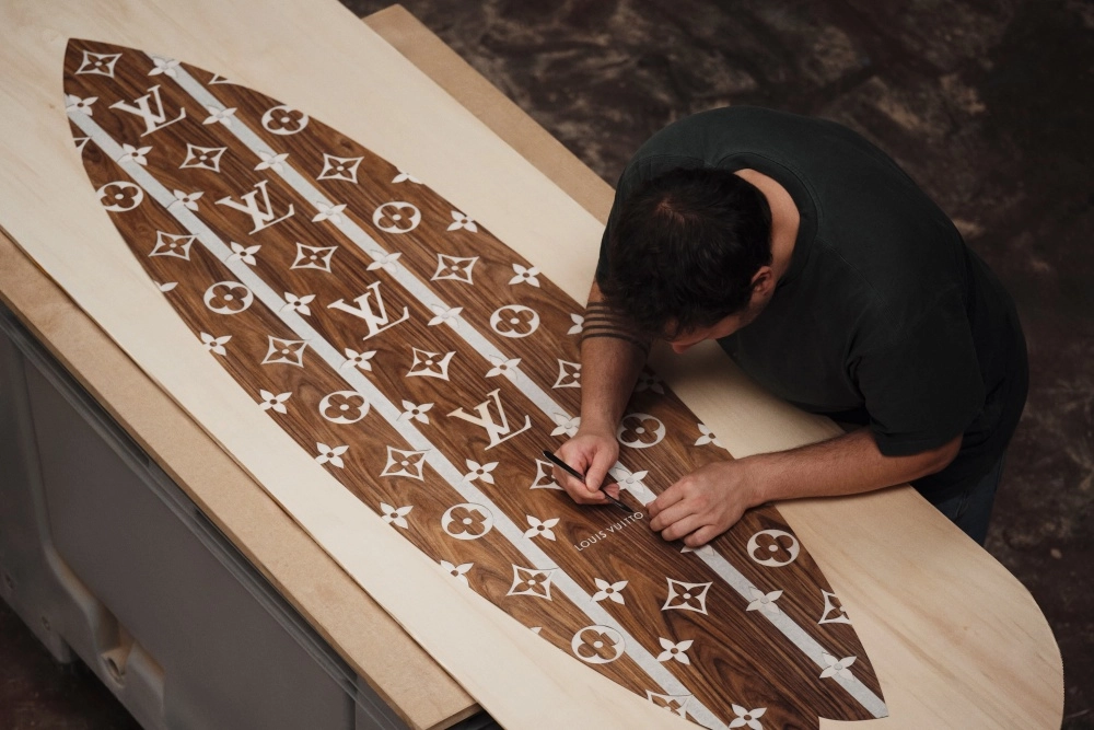 Üstten bakışla çekilen bu görselde, koyu renkli, Louis Vuitton monogramları ve beyaz çizgilerle süslenmiş ahşap bir sörf tahtası üzerinde dikkatle detay çalışması yapan bir zanaatkar, lüks markanın bu özel iş birliği koleksiyonu için ahşap işçiliği ustalığını sergiliyor.