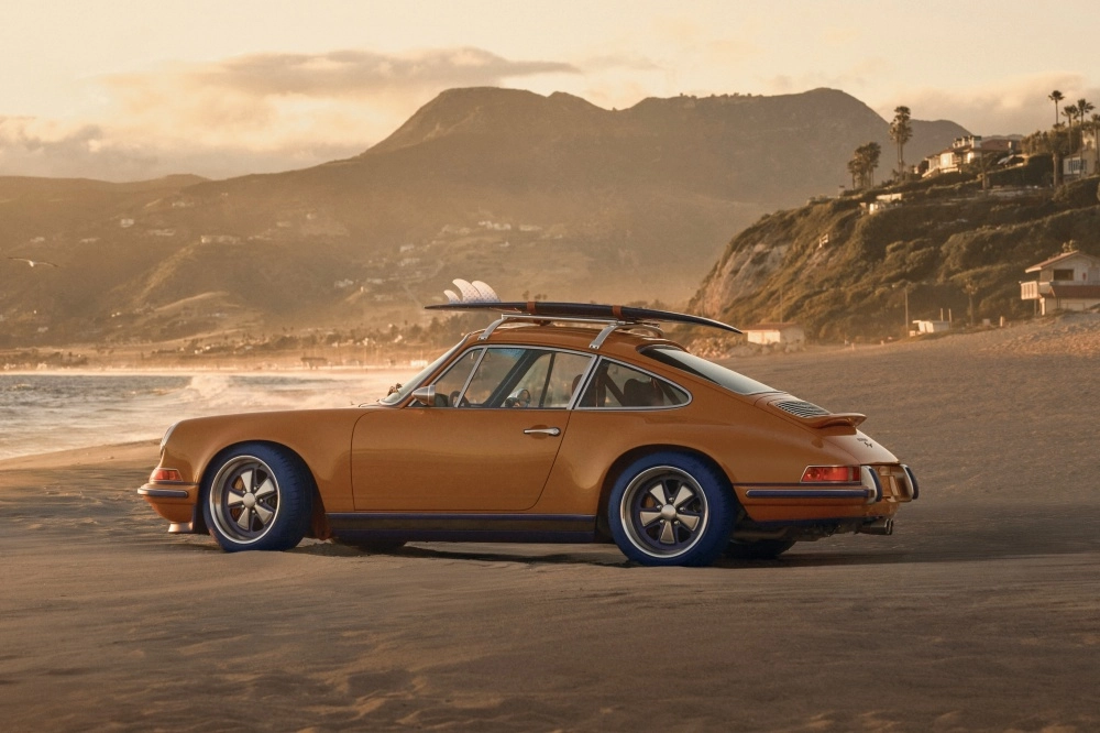 Kumlu bir sahilde, gün batımının sıcak renkleriyle aydınlanmış, tavanında sörf tahtası taşıyan, özel üretim mavi lastiklere sahip karamel tonlarında bir Singer Porsche 911 restomod otomobil, lüks bir yaşam tarzı iş birliğini yansıtıyor.