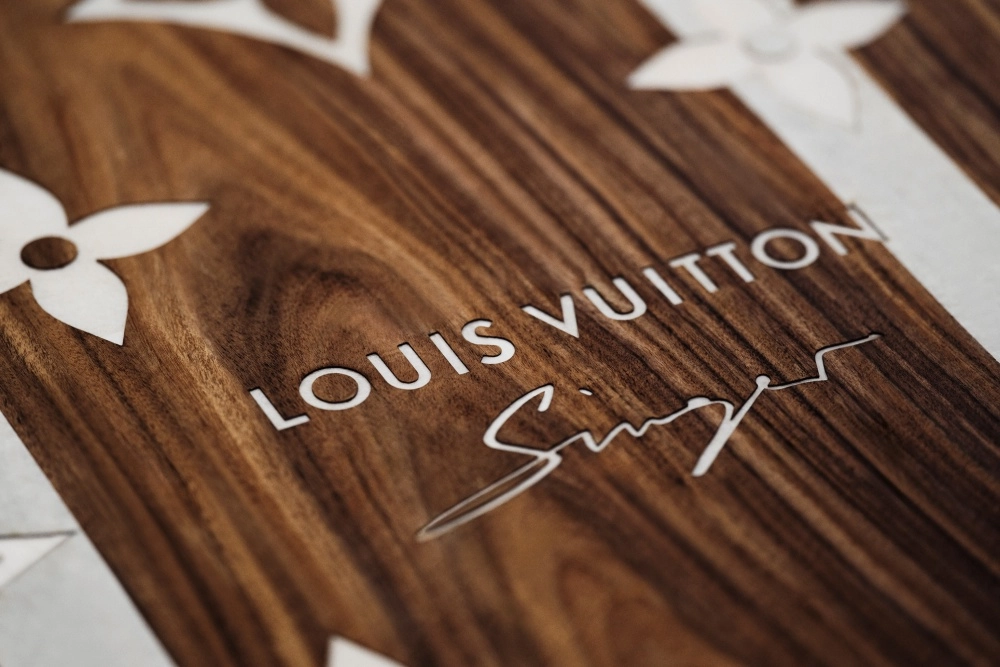 Louis Vuitton'ın ikonik monogram desenleri ve Louis Vuitton ile Singer logolarının koyu renk ahşap üzerine beyaz oymalarla işlendiği, lüks bir restomod iş birliği koleksiyonundan zarif bir tasarım detayı yakın çekimde gösteriliyor.
