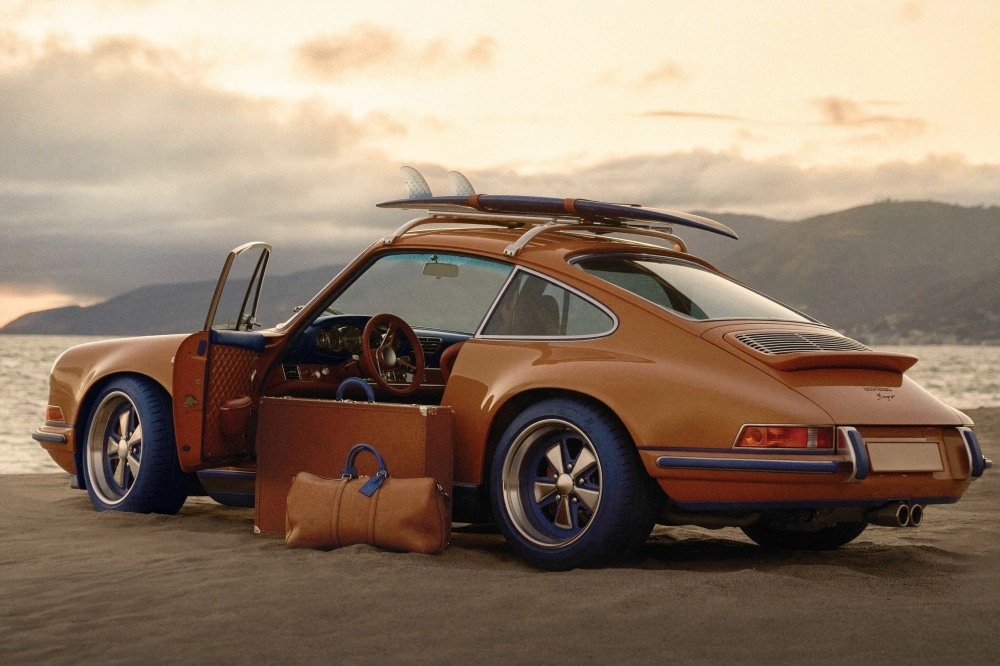 Gün batımında kumsal üzerine park etmiş, mavi detaylara ve mavi lastiklere sahip, kapısı açık kahverengi Singer Porsche 911 restomod otomobilin yanında, Louis Vuitton iş birliğiyle tasarlanmış gibi duran kahverengi valizler ve bir seyahat çantası ile tavanında sörf tahtası bulunan estetik bir koleksiyon.