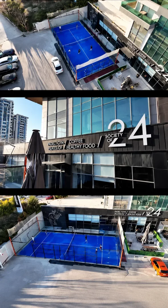 Ankara'daki modern Society of 24 spor kompleksinde, parlak mavi zemini ve cam duvarlarıyla dikkat çeken padel kortları, kortlarda aktif olarak oyun oynayan sporcular ve tesisin üzerinde Padel/Gym/Coffee/Healthy Food yazılı şık tabelasıyla birlikte dışarıdan çekilmiş geniş bir görüntü.
