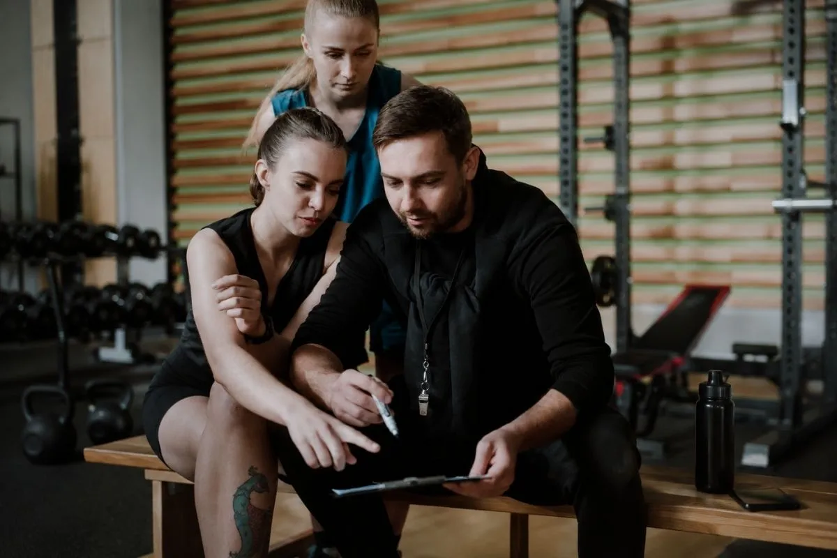 Bir spor salonunda, bir antrenör elindeki panoya işaret ederek yanındaki kadın sporcuya bir antrenman programını açıklarken, arkalarındaki başka bir kadın da ilgiyle dinliyor; bu görüntü sporda plato dönemiyle başa çıkmak ve gelişimi sürdürmek için program güncellemelerinin önemini vurguluyor.