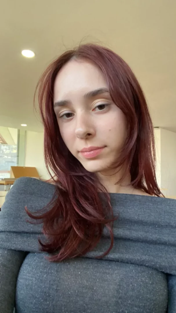 Sude Çetin'in, omuzları açık gri örgü kazağıyla, kızıl kahverengi uzun saçlarının yüzünü yumuşak bir şekilde çerçevelediği, doğal tonlarda makyajı ve hafifçe gülümseyen ifadesiyle doğrudan kameraya baktığı, sade bir arka plana sahip yakın çekim bir portre.